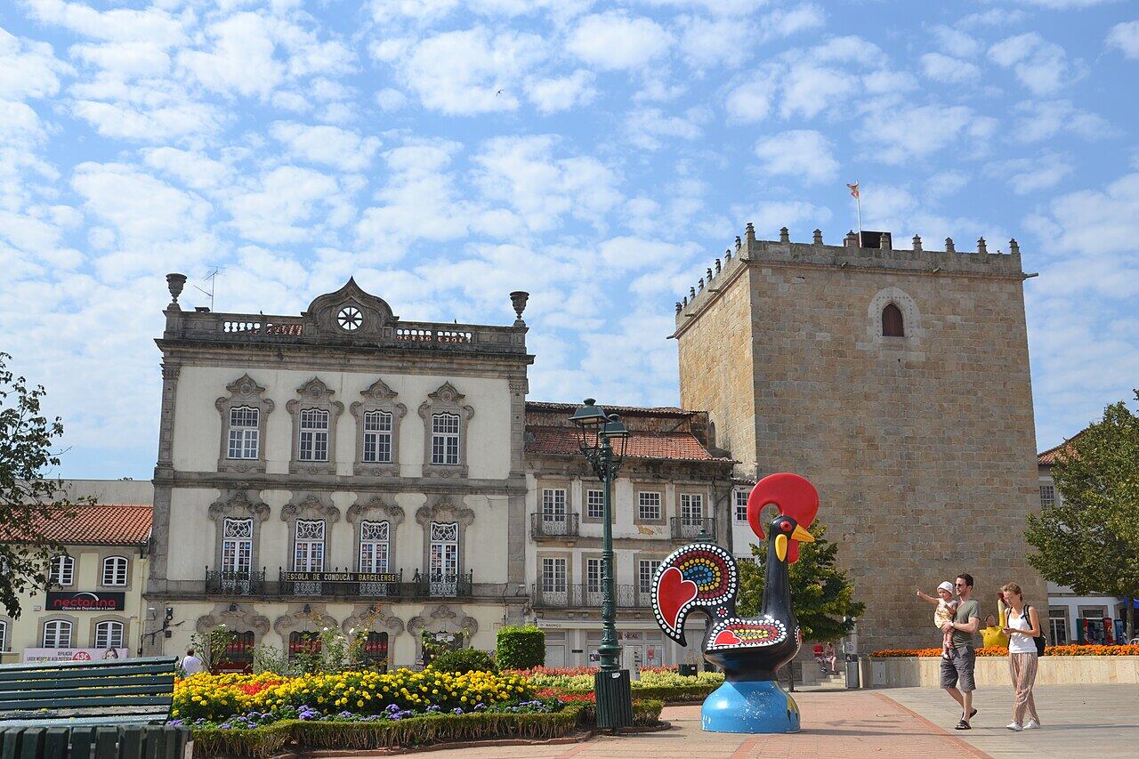 Square with a building with an ornate facade, a stone tower and a sculpture of a colourful rooster