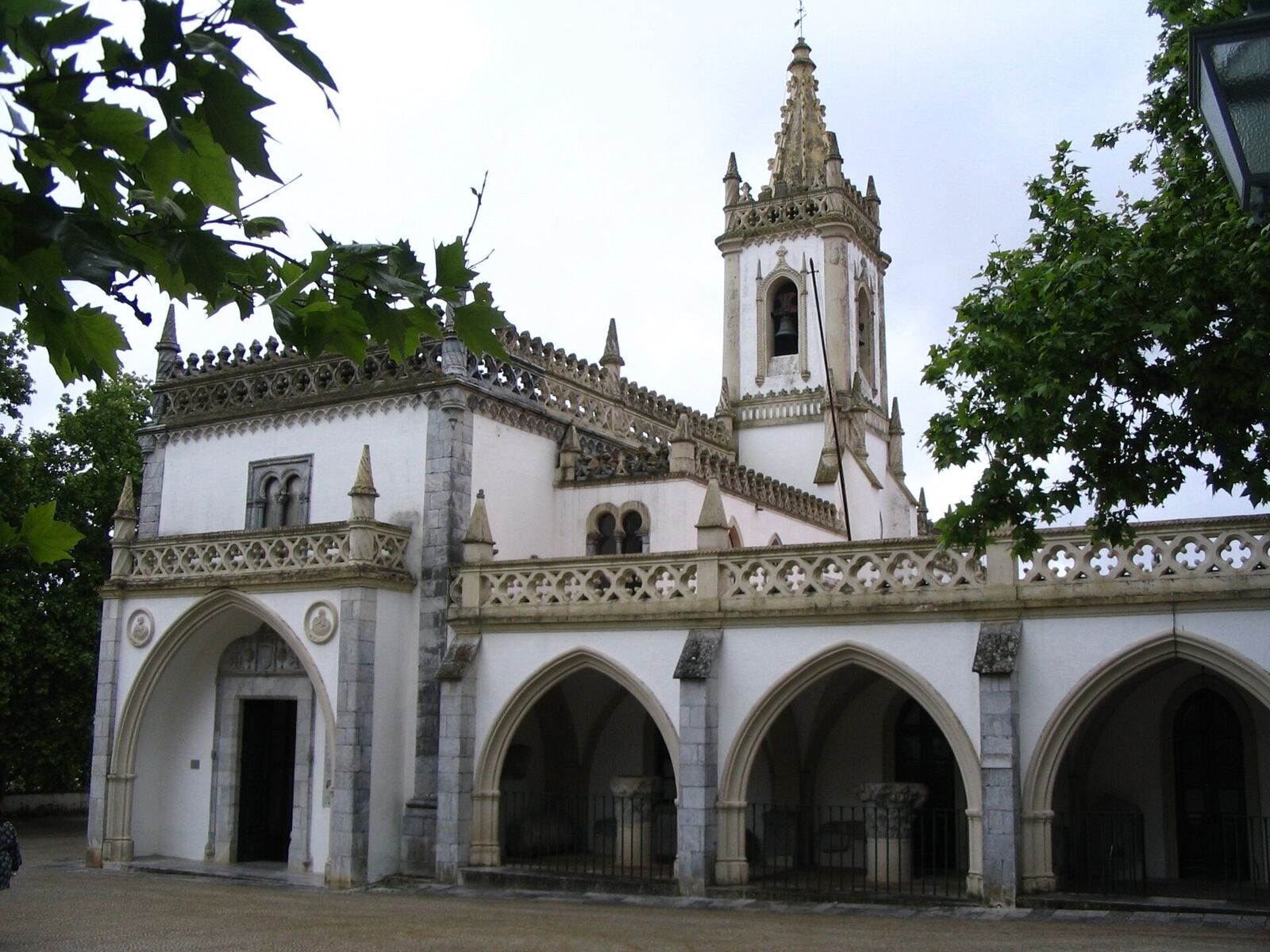 Old building of pale stone, with arcades, an ornate balcony and a bell tower, among trees