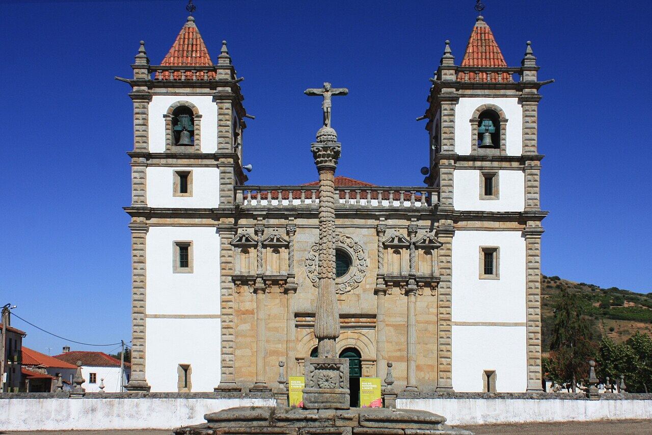 Church with two towers and a stone cross in front, under a blue sky