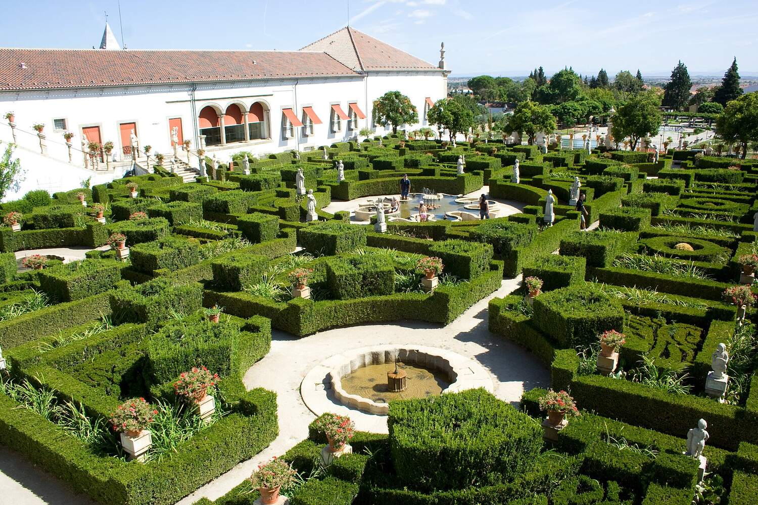 Box-hedge garden with geometric beds and statues, in front of a white building with a red roof