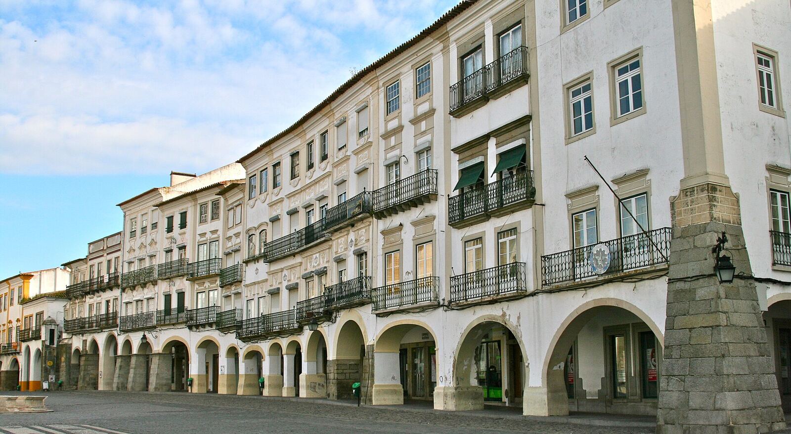 Row of white buildings with iron balconies above a stone arcade, on a square