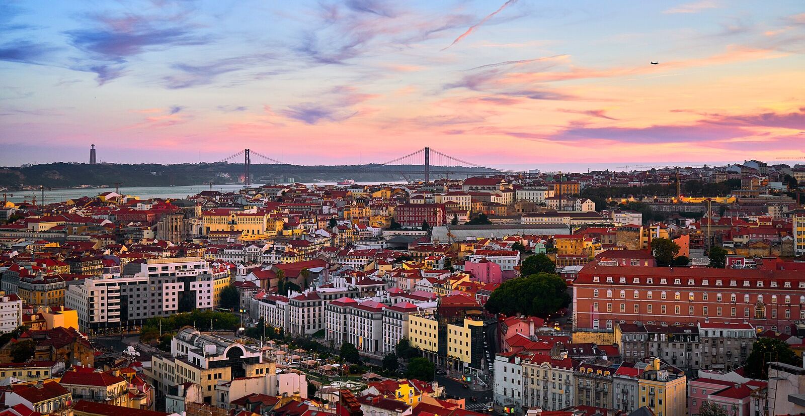 City rooftops and buildings at sunset, with the Tagus river and a bridge in the distance