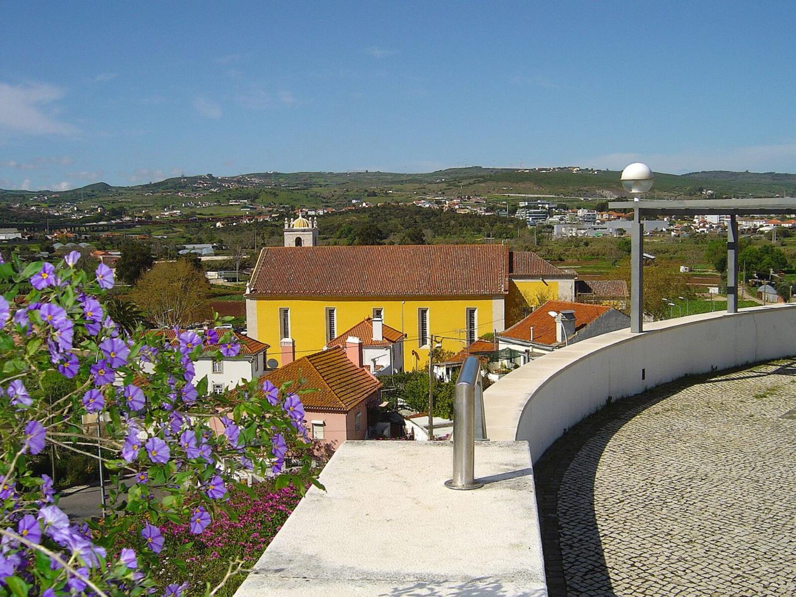 Yellow church with a red roof beside a flowery lookout, with green fields and hills behind