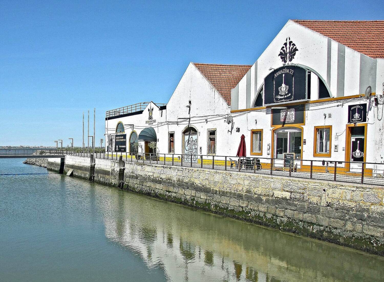 White building with a red roof and yellow trim, beside a stone quay and calm water