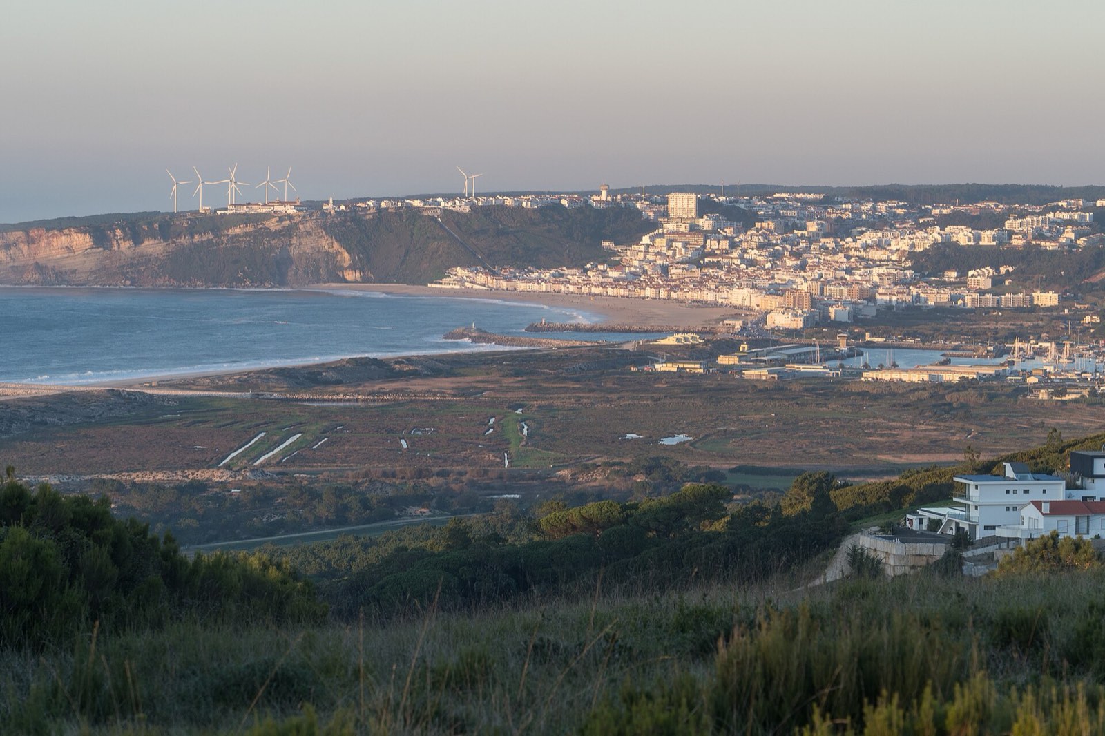 Bay with a long sandy beach, a cliff with wind turbines on top and the town of white houses behind, at the end of the day