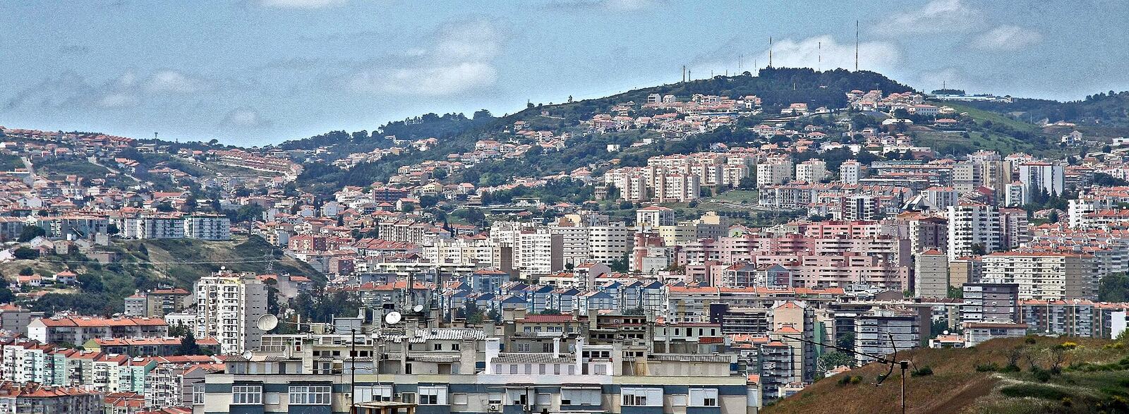 Hillside covered with apartment blocks and houses, with antennas on the crest and a blue sky