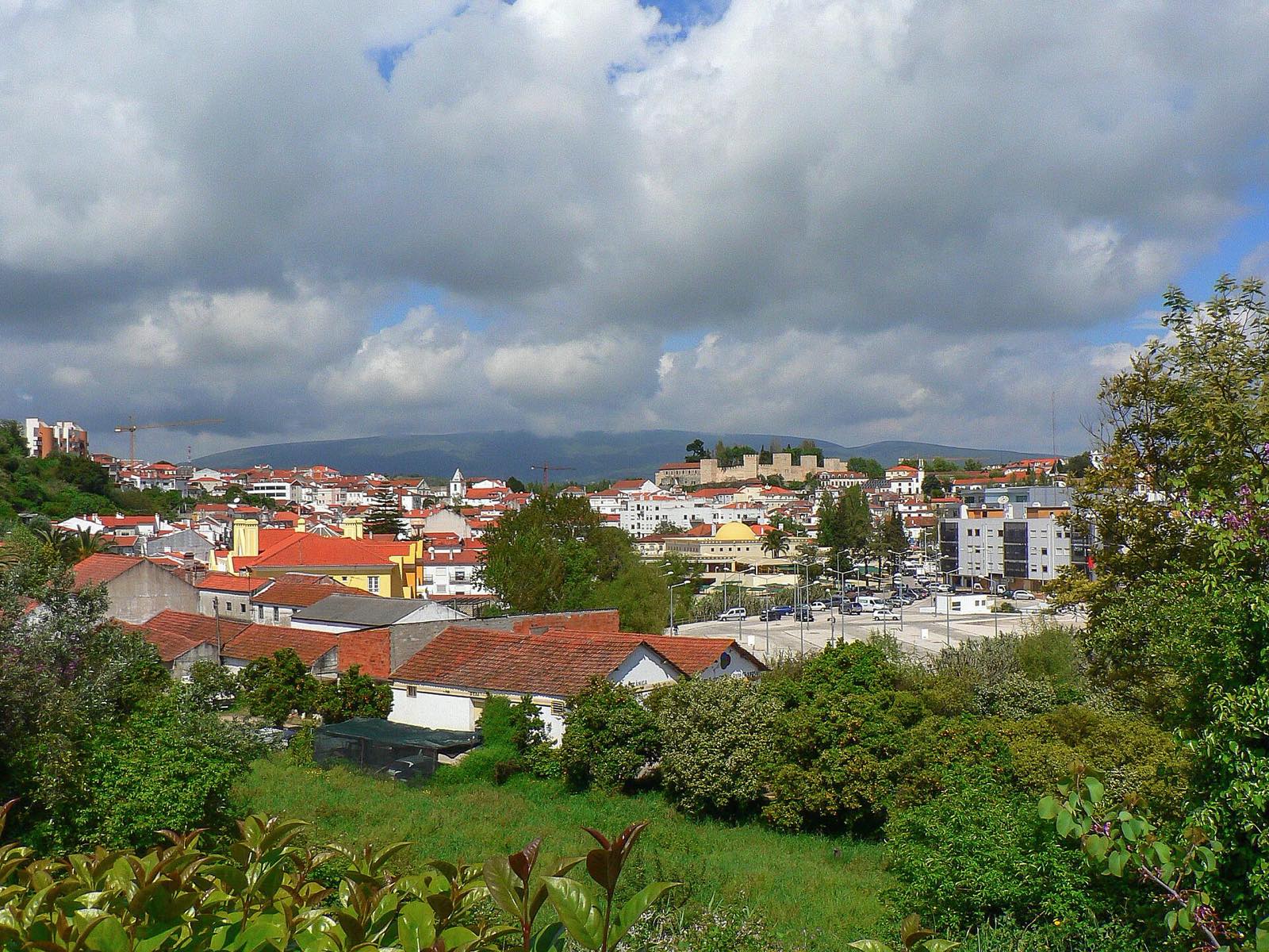Red roofs of a town, with the walls of a castle in the distance and a mountain behind, under heavy clouds